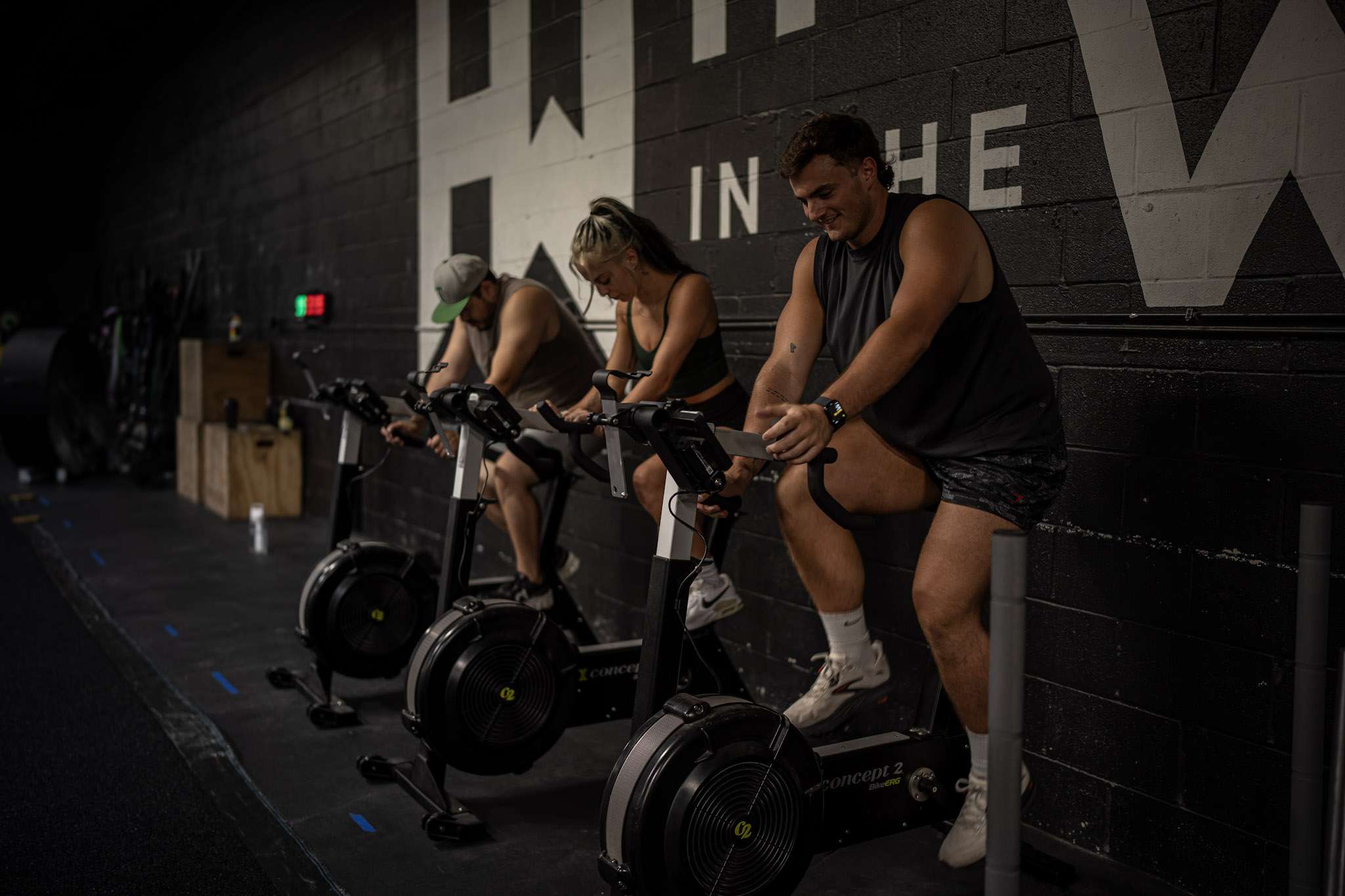 Three athletes riding Concept2 BikeErgs under the Hunger in the Wild wall logo during an engine session at our Dallas gym