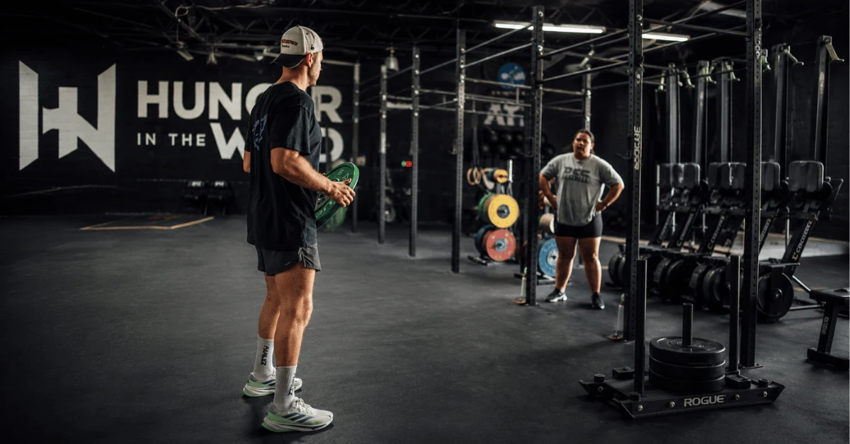 Strength Training for HYROX: Why Getting Stronger Makes You Faster on Race Day Coach instructs an athlete inside Hunger in the Wild Gym while explaining strength and conditioning techniques during a HYROX training session.