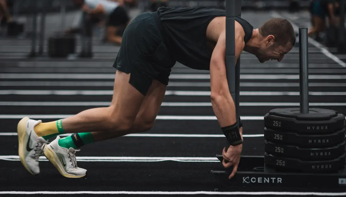 HYROX vs Spartan DEKA: What's Actually Different, and Which One Should You Race? An athlete pushes a loaded sled past stacked HYROX plates during a race.